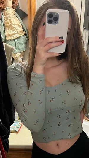 Woman in floral crop top and black pants, holding phone, mirror selfie in bedroom.