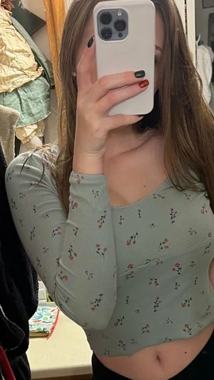Woman in floral long-sleeve top taking mirror selfie in bedroom.