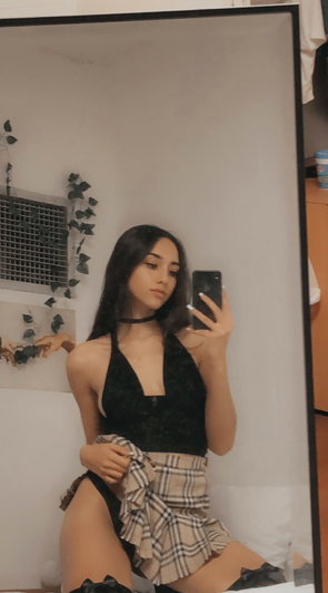 Woman in black lace top and plaid skirt takes mirror selfie in bedroom.