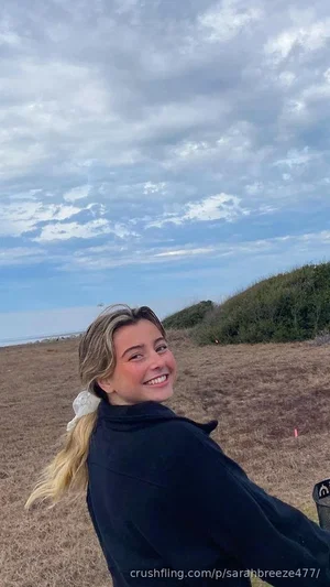 Woman with blonde hair in a ponytail, wearing a dark jacket, smiling outdoors in a grassy field.