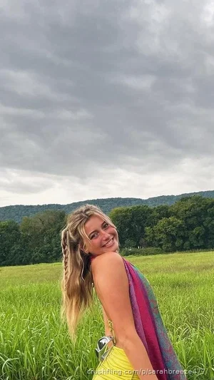 Woman with blonde braided hair in a yellow top and colorful scarf, standing in a grassy field with