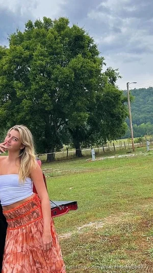 Blonde woman in strapless top and orange skirt standing outdoors near a tree and grassy field.