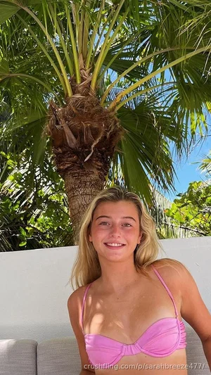 Blonde woman in pink bikini top posing outdoors near palm tree.