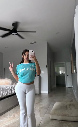 Woman in teal crop top and white pants taking mirror selfie in bedroom.