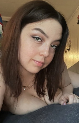 Woman with brown hair and makeup lying on bed, bare shoulders, wearing necklace.