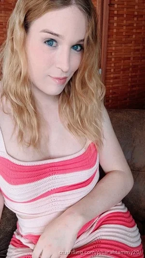 Woman with blonde hair and blue eyes wearing pink and white striped top, sitting indoors.