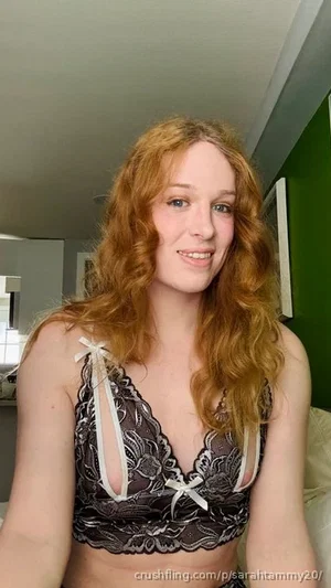 Woman with curly red hair wearing black lace bra with white bows, indoors.