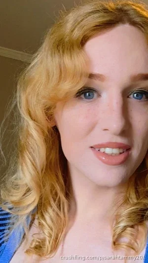 Blonde woman with blue eyes and curly hair, wearing a blue top, indoors.