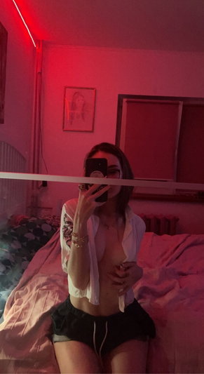 Woman in open shirt and shorts taking selfie on bed.
