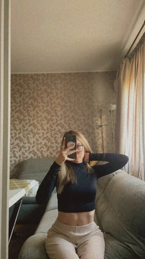 Blonde woman with glasses in crop top taking mirror selfie on couch.
