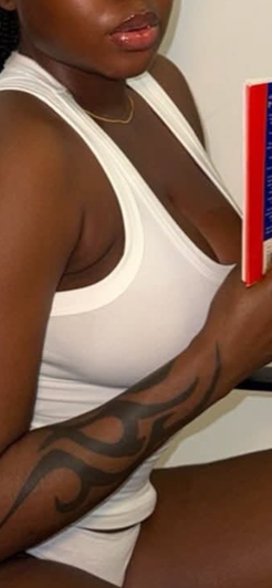 Curvy woman in white tank top with arm tattoo indoors.