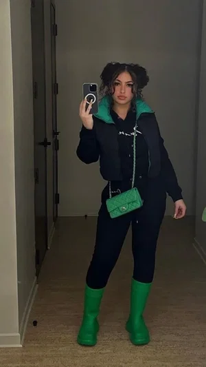 Woman in black outfit with green boots and bag, taking mirror selfie in hallway.