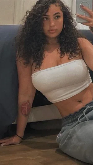 Woman with curly hair in white crop top and jeans, sitting on floor near couch.