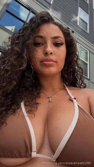 Woman with curly hair in a brown bikini top, standing outdoors near a building.