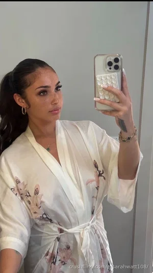 Woman in white floral robe taking mirror selfie, dark hair in ponytail, indoor setting.