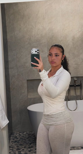 Woman in white top and gray leggings taking mirror selfie in bathroom.