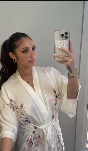 Woman in white floral robe taking mirror selfie, dark hair in ponytail, indoor setting.