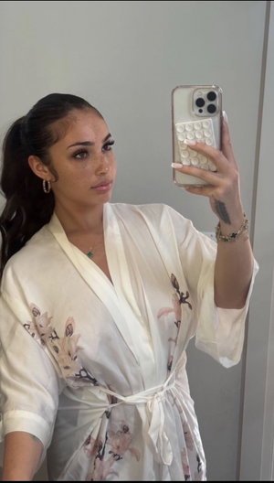 Woman in white floral robe taking mirror selfie, dark hair in ponytail, indoor setting.