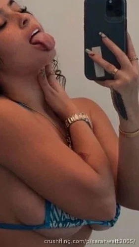 Woman in blue patterned bikini top taking mirror selfie, tongue out, hand on neck.