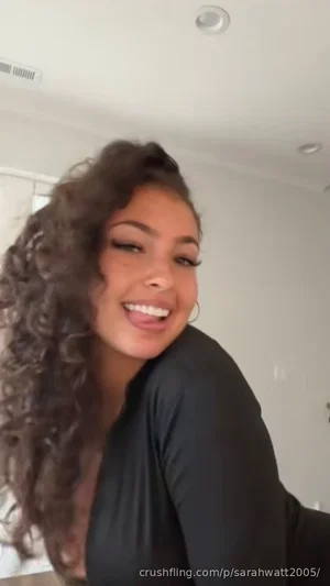 Woman with curly hair in black top, smiling indoors.