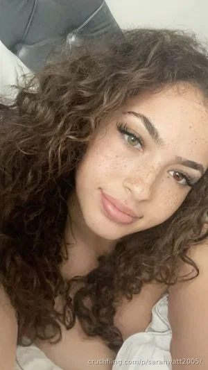 Woman with curly hair and freckles, wearing white top, lying down indoors.
