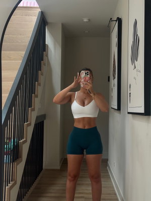 Woman in white crop top and teal shorts taking mirror selfie in hallway.