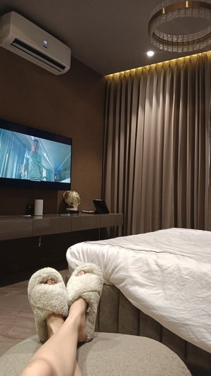 Person's feet in fluffy slippers on ottoman, watching TV in bedroom.