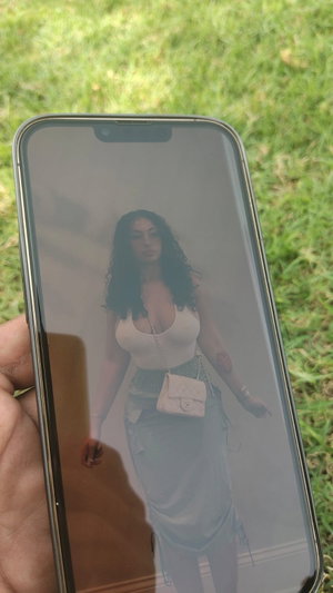 Woman with long dark hair in white top and light skirt, standing outdoors on grass.
