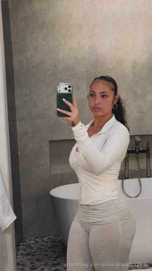 Woman in white long-sleeve top and gray leggings taking mirror selfie in bathroom with bathtub.