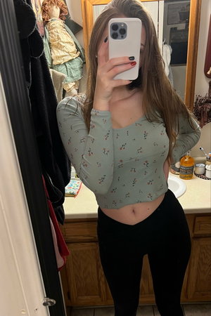 Woman taking mirror selfie in bathroom wearing floral crop top.