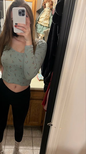 Woman in floral crop top and black leggings taking mirror selfie in bathroom.