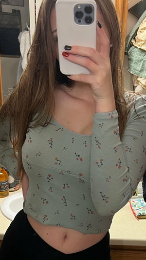 Woman with long brown hair in floral top and black bottoms, taking mirror selfie in bathroom.