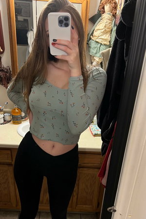 Woman in floral crop top and black leggings taking mirror selfie in bathroom.