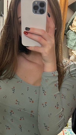 Woman with long brown hair in floral long-sleeve top taking mirror selfie indoors.