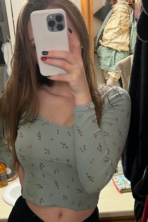 Woman with long brown hair in floral crop top and black bottoms, taking mirror selfie in bathroom.