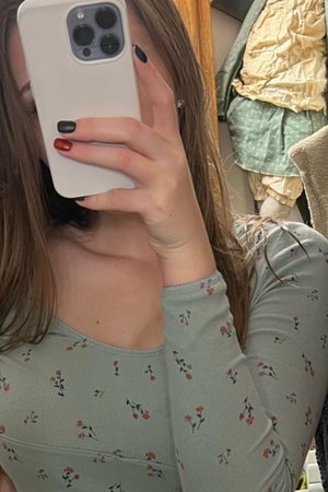 Woman with long hair in floral top taking mirror selfie indoors.