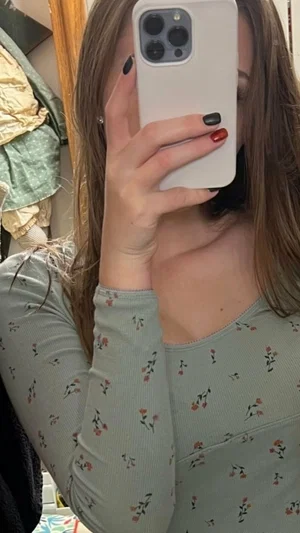 Woman in floral long-sleeve top taking mirror selfie, hair covering face partially.