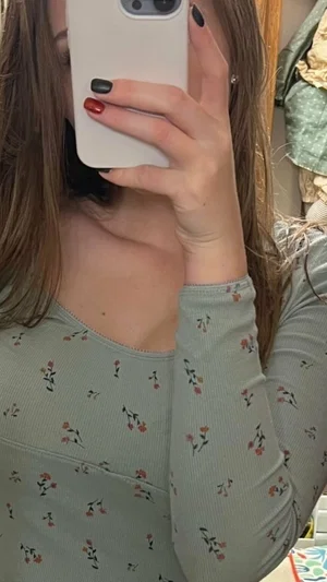 Woman with long brown hair in floral long-sleeve top taking mirror selfie indoors.