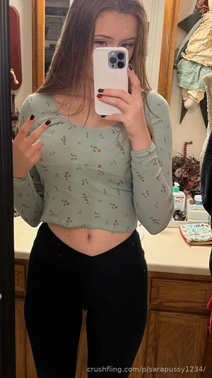 Woman in floral crop top and black leggings taking mirror selfie in bathroom.