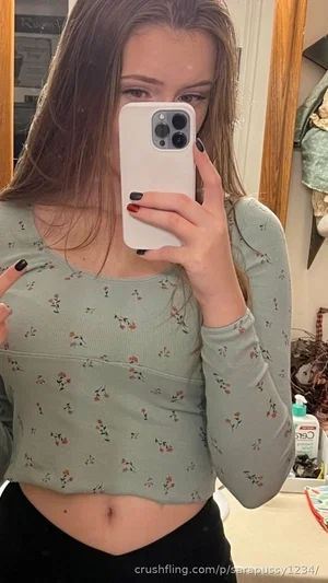 Woman in floral crop top and black pants taking mirror selfie in bedroom.