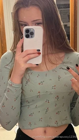 Woman in floral crop top and black pants taking mirror selfie indoors.