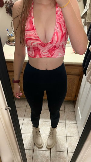 Woman in pink crop top and black leggings taking bathroom mirror selfie.