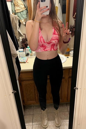 Curvy woman in pink top and black leggings taking bathroom mirror selfie.