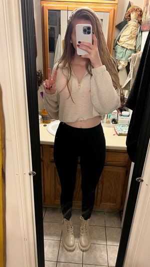 Brunette woman taking mirror selfie in bathroom wearing crop sweater.
