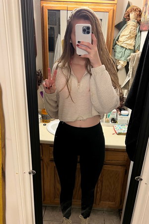 Woman with long hair taking mirror selfie in bathroom wearing sweater and leggings.