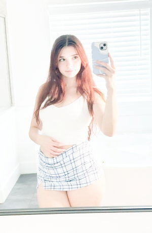 Redhead woman taking mirror selfie in white top and plaid skirt.