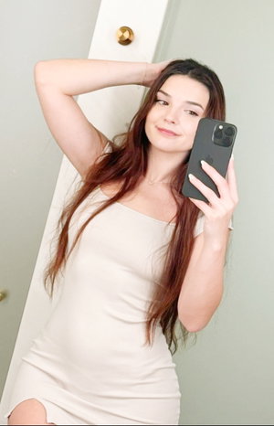 Long-haired woman in beige dress taking mirror selfie indoors.