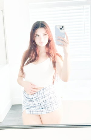 Woman with long hair in white crop top and plaid skirt taking mirror selfie indoors.