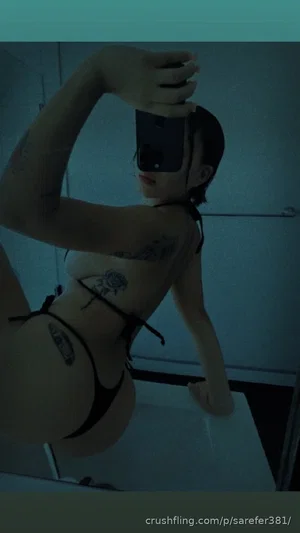 Woman in black lingerie with tattoos taking mirror selfie in bathroom.