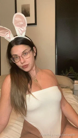 Woman with long dark hair wearing white bunny ears and a white one-piece bodysuit, sitting on a bed